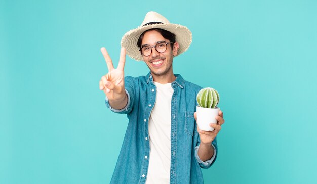 giovane ispanico che sorride e sembra felice, gesticola vittoria o pace e tiene in mano un cactus