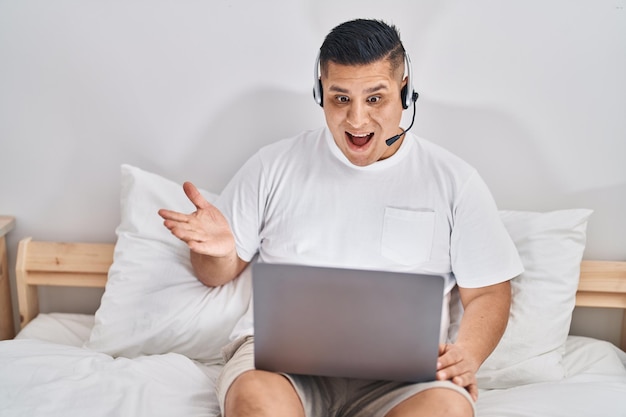 Giovane ispanico che utilizza il computer portatile sul letto che celebra il successo con un sorriso felice e l'espressione del vincitore con la mano alzata
