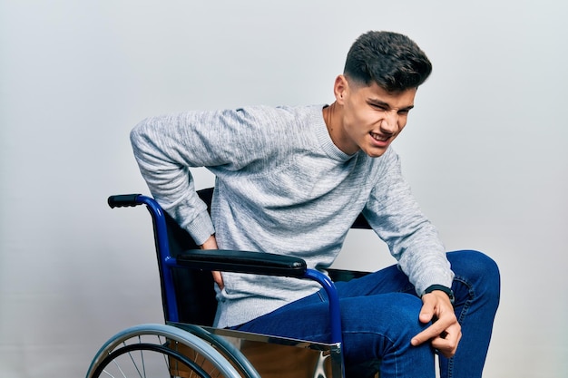 Giovane ispanico seduto su una sedia a rotelle che soffre di mal di schiena, toccando la schiena con la mano, dolore muscolare