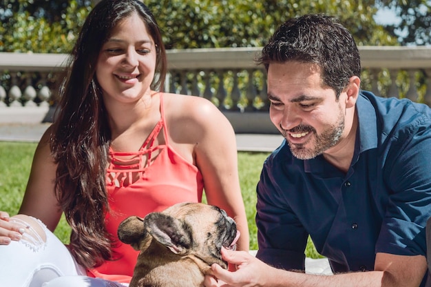 Giovane latino barbuto che gioca con il suo bulldog francese nel parco con la sua ragazza