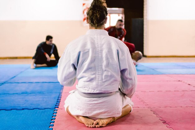 Giovane latino e maschio ispanico con i capelli lunghi seduto sulle gambe su un pavimento tatami guardando una lotta di jiujitsu