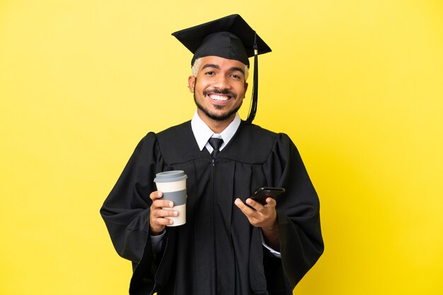 Giovane laureato uomo colombiano isolato su sfondo giallo tenendo il caffè da asporto e un mobile