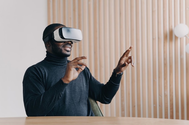 Giovane lavoratore africano gioioso uomo d'ufficio in occhiali per cuffie VR che gioca al gioco 3D preferito