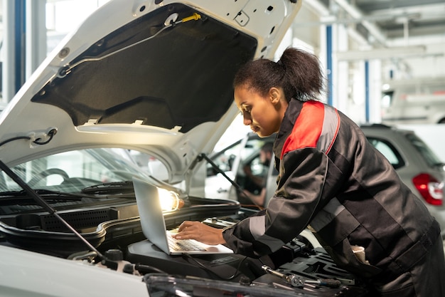 Giovane lavoratore del servizio di manutenzione dell'auto che si china sul vano motore aperto mentre utilizza il laptop per consultare fonti online