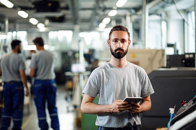 Giovane lavoratore manuale con tavoletta digitale in piedi in una struttura industriale e guardando la fotocamera