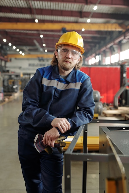 Giovane lavoratore maschio barbuto di un grande stabilimento o fabbrica o caposquadra in abiti da lavoro e casco protettivo appoggiato a attrezzature industriali