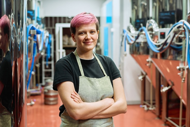 Giovane lavoratrice bionda sorridente della fabbrica di produzione di birra in abiti da lavoro incrociando le braccia per il petto mentre si trova contro l'attrezzatura di lavorazione