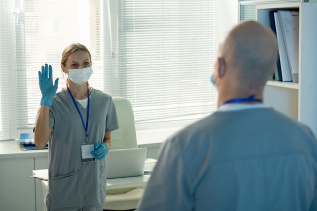 Giovane lavoratrice del laboratorio medico in abiti da lavoro protettivi agitando la mano al collega maschio calvo mentre lo saluta al mattino