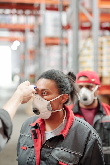 Giovane lavoratrice di magazzino in respiratore e abbigliamento da lavoro con la sua temperatura misurata con la coda di altri lavoratori dietro