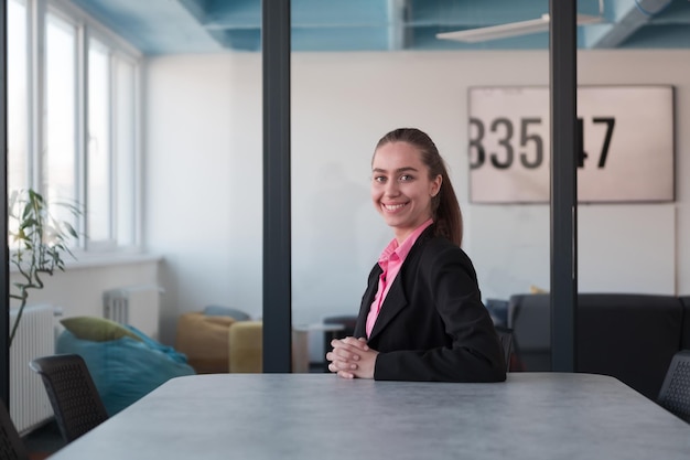 Giovane leader femminile di successo in giacca e cravatta con una camicia rosa seduta in un moderno ufficio di vetro con un sorriso determinato. Concetto di business e successo dei giovani nelle moderne attività online