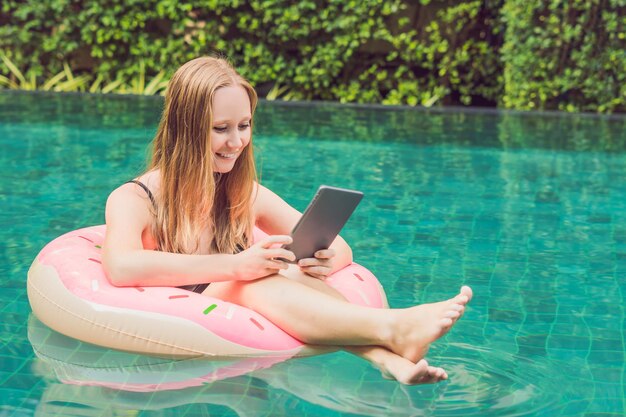 Giovane libera professionista seduta vicino alla piscina con il suo computer portatile in hotel navigando nel suo smartphone. Impegnato durante le vacanze. Concetto di lavoro a distanza. Copia spazio per il tuo testo.