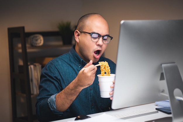 Giovane libero professionista asiatico che lavora a tarda notte Ha molto fame e mangia spaghetti istantanei caldi e lavora con il computer nell'ufficio buio di notte