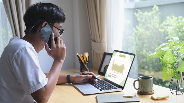 Giovane libero professionista maschio che ha una conversazione sul cellulare e lavora con un computer portatile a casa