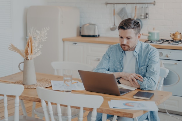 Giovane libero professionista serio e concentrato che lavora al computer portatile mentre è seduto al tavolo della cucina con le mani appoggiate su di esso, grafici e statistiche sparsi in giro, taccuino aperto accanto a lui