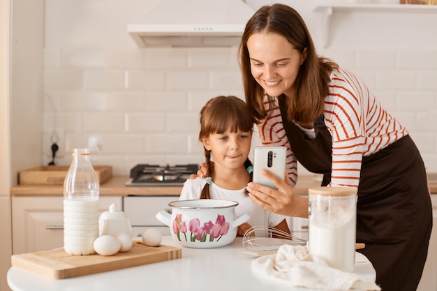 Giovane madre adulta che cucina insieme a sua figlia, seduta a tavola in cucina e trova la ricetta in internet tramite telefono cellulare o si fa selfie mentre cucina, la gente sorride felicemente.