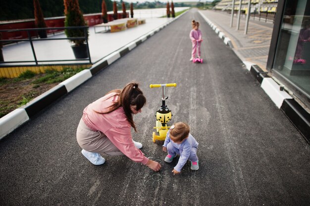 Giovane madre alla moda con due ragazze all'aperto. La famiglia sportiva trascorre il tempo libero all'aperto con gli scooter. Dipinto con gesso sull'asfalto.