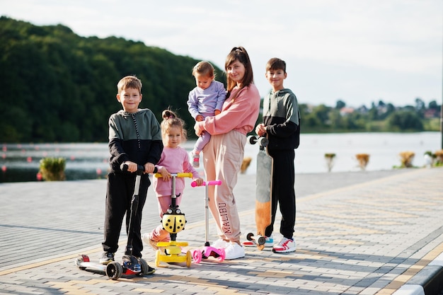 Giovane madre alla moda con quattro bambini all'aperto La famiglia sportiva trascorre il tempo libero all'aperto con scooter e pattini