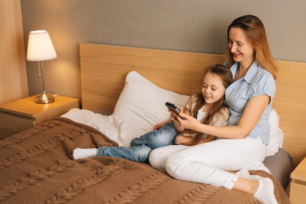 Giovane madre allegra che abbraccia alla figlia sorridente sveglia insieme guardando video divertenti sul telefono cellulare