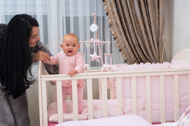 Giovane madre amorevole che aiuta la sua piccola figlia sorridente a stare nella sua culla in un asilo nido