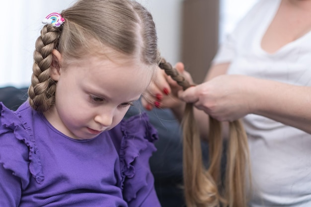 Giovane madre amorevole che spazzola i capelli delle figlie del bambino e acconcia l'acconciatura della treccia della pettinatura a casa