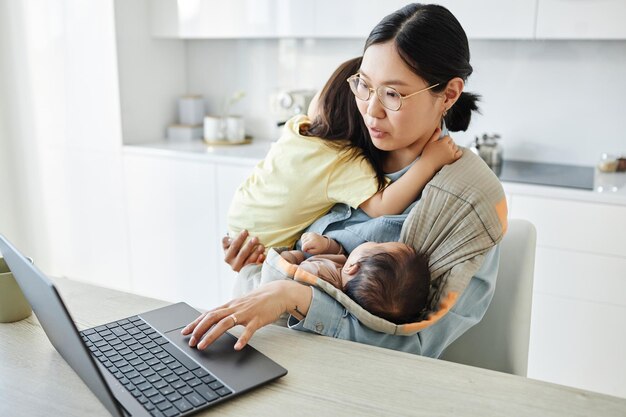Giovane madre asiatica con due bambini che lavorano online sul computer portatile mentre si siede in cucina