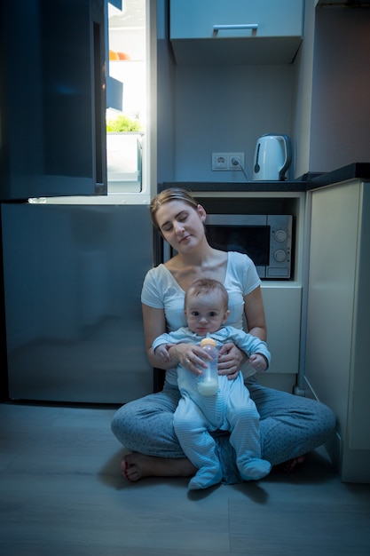 Giovane madre assonnata seduta in cucina con suo figlio accanto al frigorifero. Concetto di alimentazione del bambino di notte