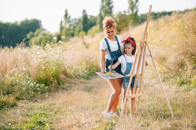 Giovane madre attraente insegna figlia a dipingere nel parco estivo. Attività all'aperto per il concetto di bambini in età scolare