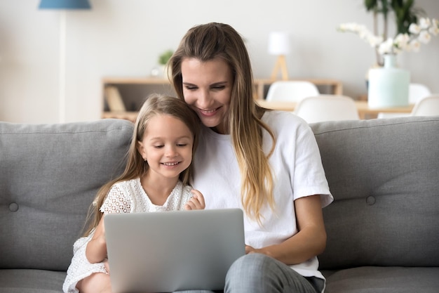 Giovane madre caucasica seduta sul divano rilassarsi con la piccola figlia in età prescolare guardare video divertenti sul portatile insieme sorridendo madre o babysitter riposo con bambina piccola usando moderno computer gadget a casa