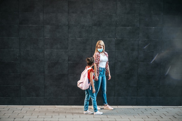 Giovane madre che cammina con la sua piccola figlia sulla strada della città. Indossano maschere protettive per il viso. Ritorno a scuola e nuovo concetto di stile di vita del coronavirus.