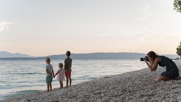 Giovane madre che scatta foto dei suoi tre bambini, due ragazzi e una ragazza, mentre stanno sulla spiaggia la sera tenendosi per mano