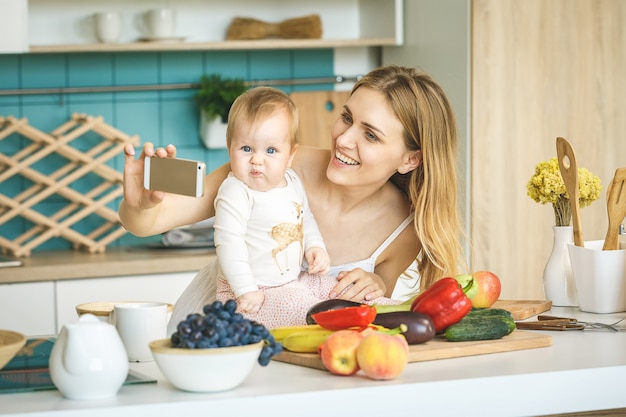 Giovane madre che sorride, che cucina e che gioca con sua figlia in una cucina moderna. Lavorare a casa. Donna con un bambino che fa un selfie.