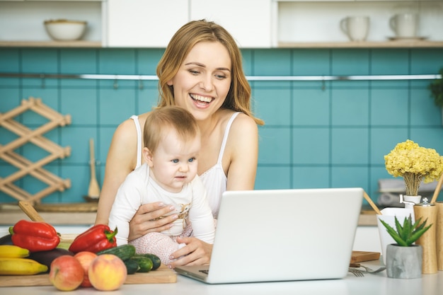 Giovane madre che sorride, che cucina e che gioca con sua figlia in una cucina moderna. Usando il telefono.