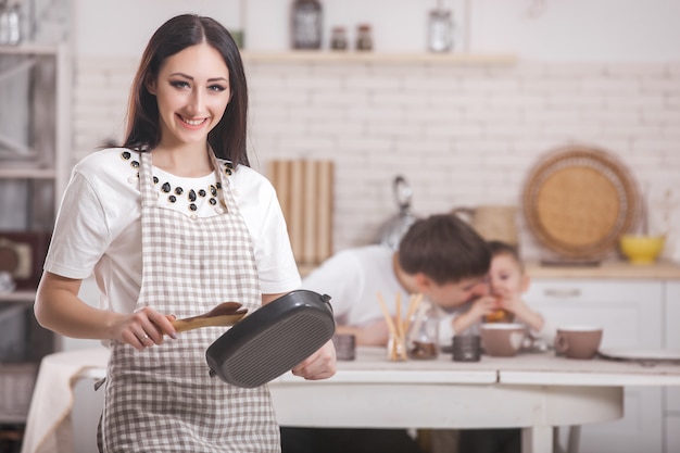 Giovane madre che sta davanti alla sua famiglia sulla cucina. Famiglia felice cenando o facendo colazione. Donna che prepara la cena per il marito e il bambino piccolo.