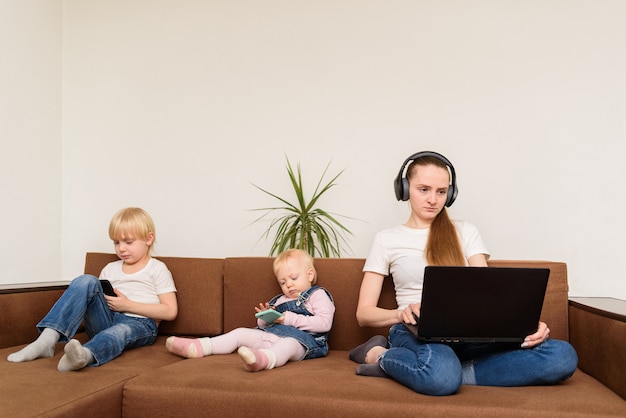 Giovane madre con il computer in mano e bambini con telefoni seduti sul divano. Uso eccessivo di dispositivi