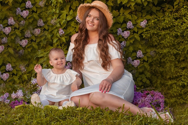 giovane madre dai capelli castani in prendisole bianco con una piccola figlia seduta da un picnic di un cespuglio di lillà