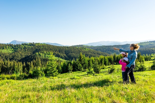 Giovane madre e due piccole figlie viaggiatori stanno su un pendio con una splendida vista sulle colline ricoperte da una fitta foresta di abeti contro il cielo blu in una calda giornata estiva di sole