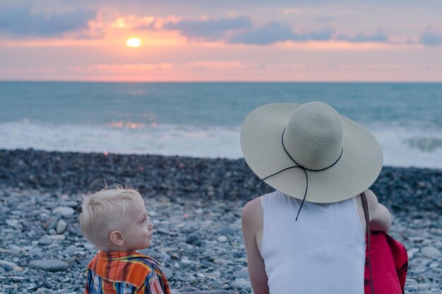 Giovane madre e figlio seduti sulla spiaggia contro il mare e il tramonto Concetto di madre single