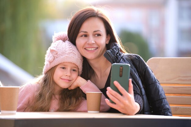 Giovane madre e sua figlia che scattano foto con la fotocamera selfie del telefono seduti al caffè di strada con bevande calde in una giornata di sole autunnale Presenza sui social media nel concetto di vita quotidiana