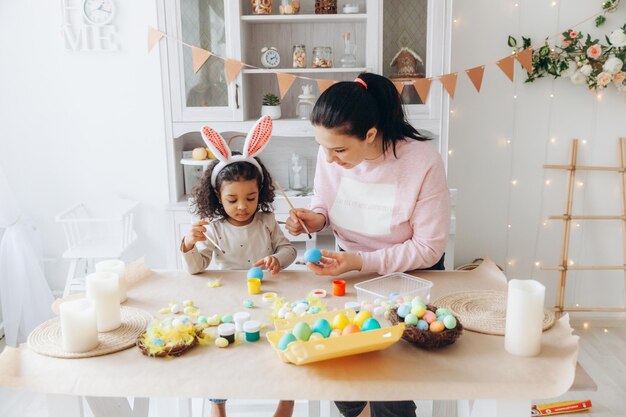 Giovane madre europea che insegna al bambino afroamericano come dipingere e decorare le uova con le vernici per le vacanze di pasqua seduti insieme al tavolo della cucina