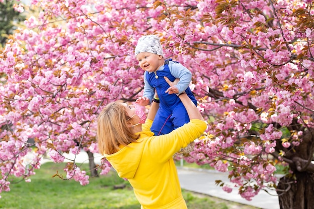 Giovane madre in maschera medica con adorabile figlioletto nel parco con albero di fiori di ciliegio Sakura Madre e bambino felici