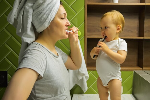 Giovane madre in turbante asciugamano e bambina lavarsi i denti nel bagno dell'appartamento Concetto quotidiano di cura orale Il bambino si rallegra in piedi sul lavandino