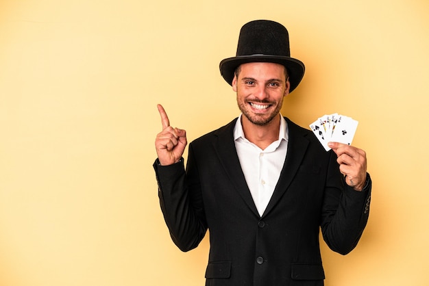 Giovane mago caucasico che tiene in mano una carta magica isolata su sfondo giallo sorridendo e indicando da parte, mostrando qualcosa in uno spazio vuoto.