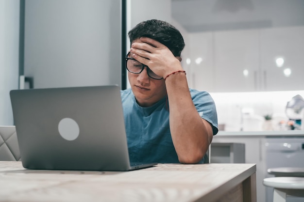 Giovane malato che si sente malsano a casa lavorando con il computer portatile sul tavolo da solo Ragazzo adolescente che tocca la testa dopo una giornata stanca Concetto di superlavoro