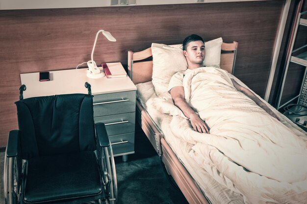 Giovane malato di sesso maschile che giace nel letto d'ospedale coperto di trapunta in corsia d'ospedale. Concetto di assistenza sanitaria