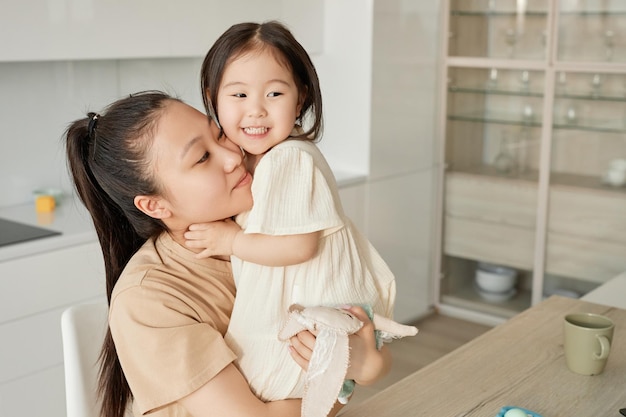 Giovane mamma asiatica che abbraccia la sua piccola figlia mentre si siedono in cucina