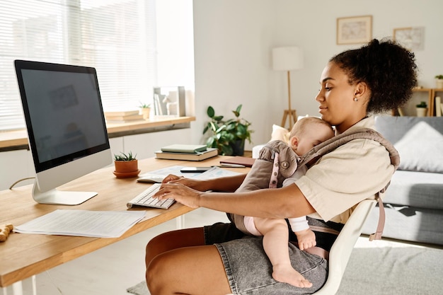 Giovane mamma seduta sul posto di lavoro davanti al monitor e digitando con il suo bambino in fionda