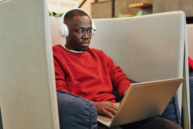 Giovane manager di ufficio maschio africano o designer in cuffia che guarda il display del laptop sulle ginocchia mentre prepara la presentazione