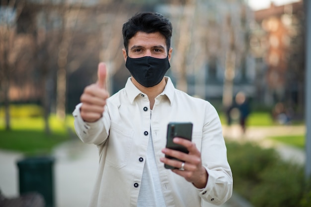 Giovane mascherato che dà i pollici in su mentre utilizza il concetto di smartphone, covid e coronavirus