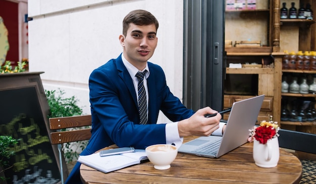 Giovane maschio allegro in tuta che punta allo schermo del laptop e guarda la fotocamera mentre spiega la strategia aziendale al tavolo nell'accogliente caffè all'aperto