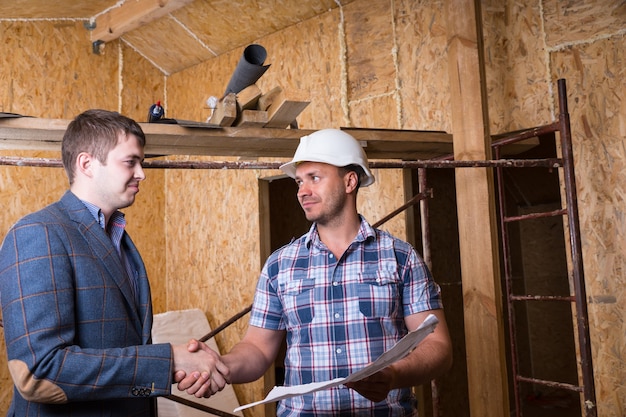 Giovane maschio architetto e lavoratore edile Foreman con piani di costruzione stringono la mano all'interno di una casa incompiuta con pannelli di compensato di particelle esposte